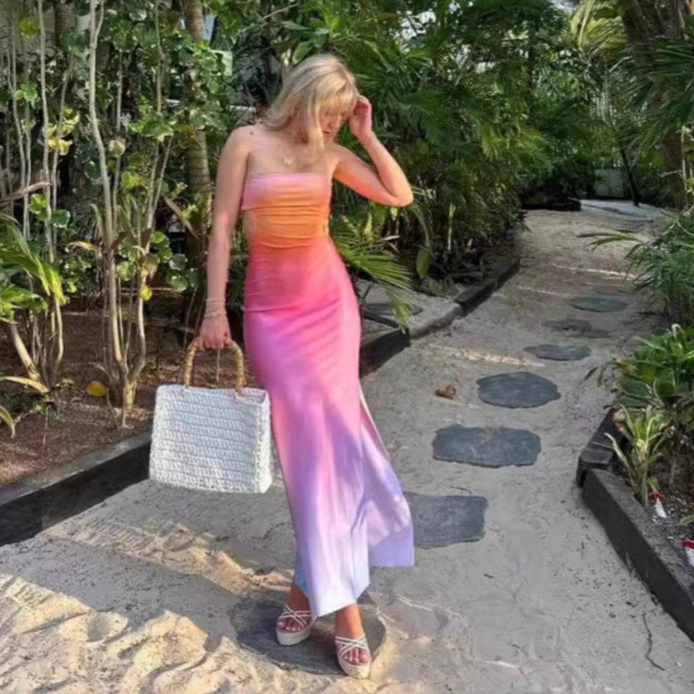 Model in a beautiful ombre strapless floor-length dress with a soft color gradient from warm orange at the bust through pink to cool purple at the hem. Full-body pose on a sunny garden stone pathway with dense green foliage, holding a woven tote bag and touching her hair.