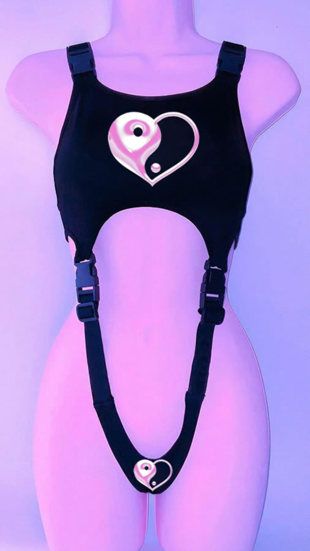Black harness-style crop top on a mannequin with adjustable buckle straps and large side cut-outs. Features a bold yin-yang heart graphic in white and pink on the chest and a smaller matching heart design at the bottom, displayed under purple lighting.