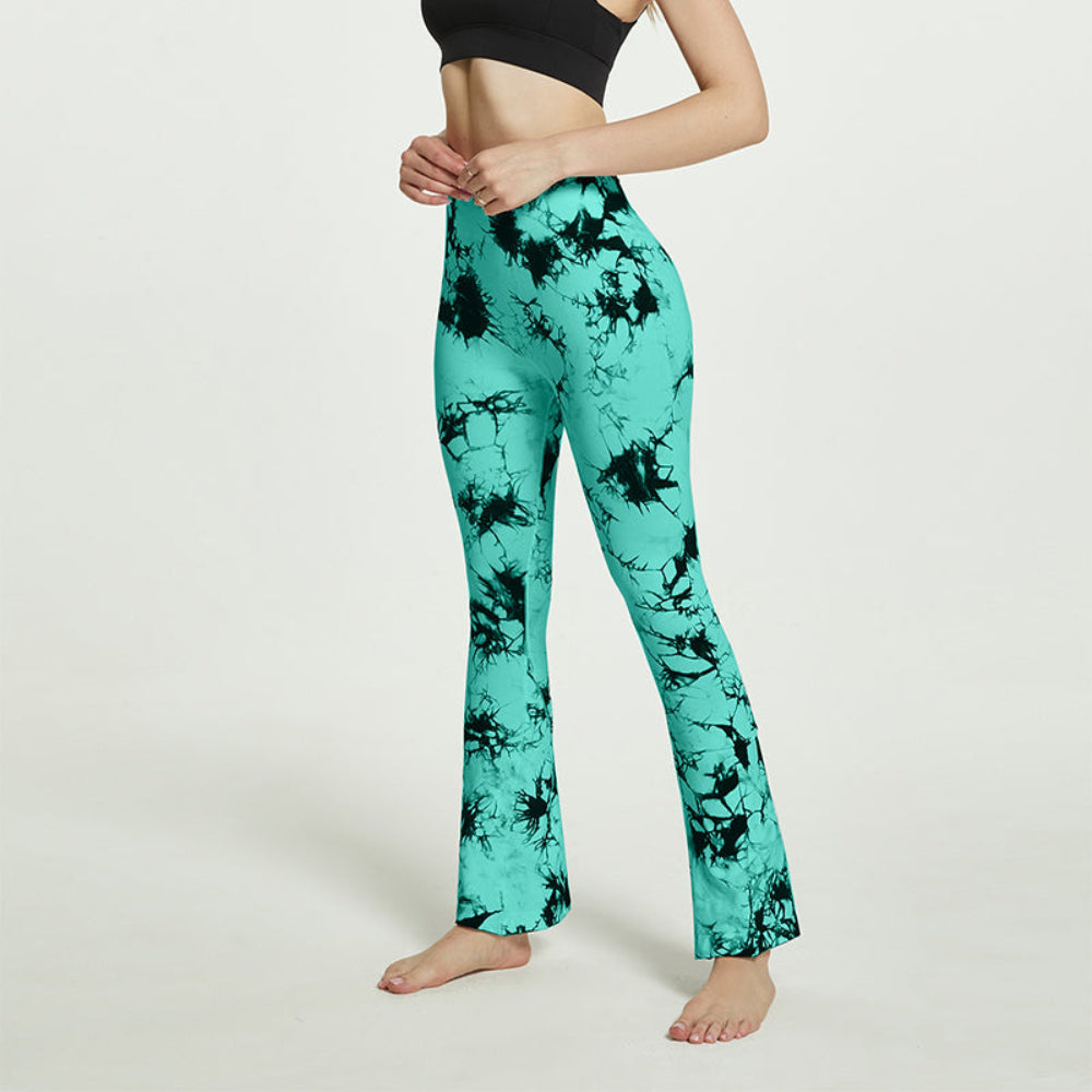 Woman modeling teal and black tie-dye high-waisted flared pants, shown from a side view while adjusting the waistband, barefoot against a clean white studio background.