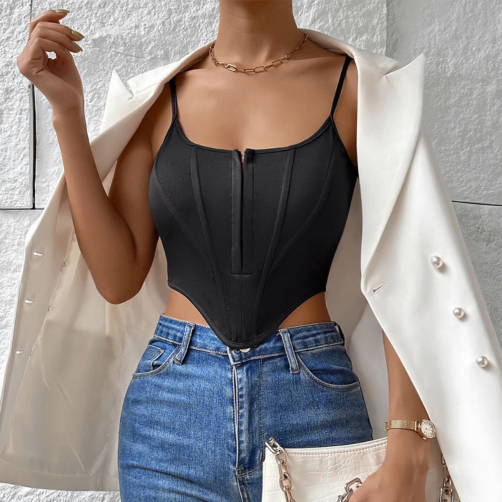 Woman wearing a black corset-style bodysuit with front zipper under an open white blazer with pearl buttons. Styled with blue denim jeans, gold chain necklace, watch, and white handbag, posing confidently with one hand near her face against a textured white wall.