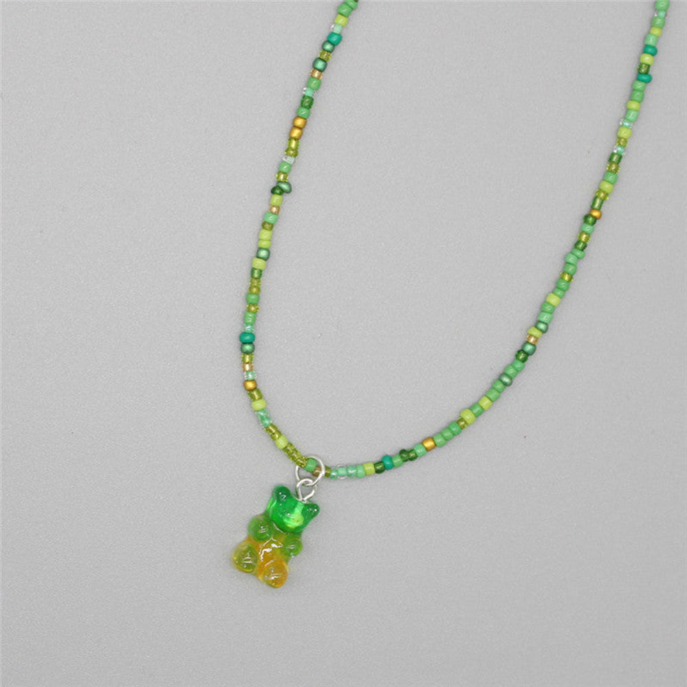 Green beaded necklace with a vibrant mix of lime, emerald, and metallic green seed beads paired with a translucent green and yellow gummy bear pendant. Playful single-strand design laid flat on a light gray background, showcasing its fresh and whimsical summer style.