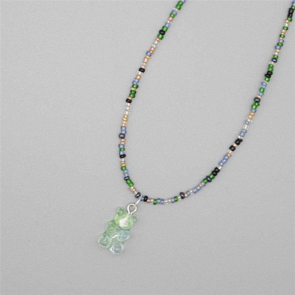 Playful beaded necklace with a mix of vibrant green, blue, black, white, and metallic seed beads. Features a translucent light green gummy bear pendant. The fun single-strand necklace is displayed flat on a light gray background, highlighting its colorful and whimsical design.