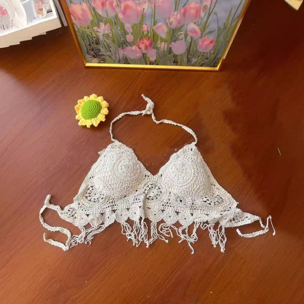 A white handmade crocheted bikini top with delicate lace patterns, triangle-style cups, halter neck ties, and a fringed hem, laid flat on a glossy wooden table. A small yellow crocheted sunflower with a green center sits to the left of the top. In the background, a framed painting of pink tulips is visible.
