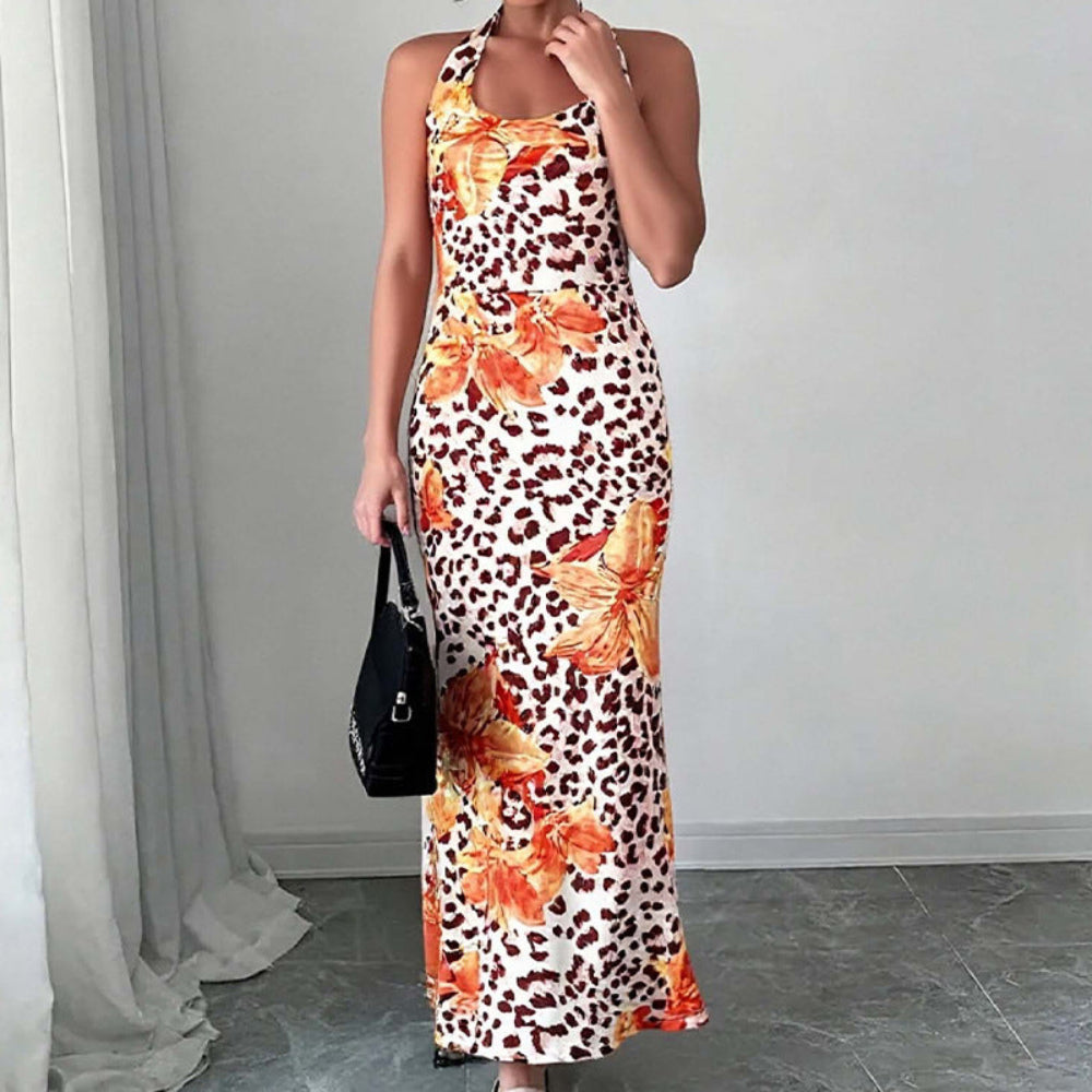 Model wearing a form-fitting sleeveless leopard-print maxi dress featuring bold orange and yellow floral overlays. Full-body pose with one hand near her neck, accessorized with a black handbag and neutral heels, in a minimalist room with white walls and curtains.
