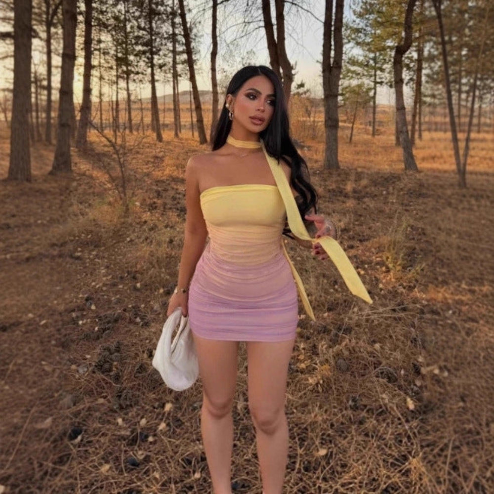 "Woman with long dark hair wearing a gradient yellow-to-pink strapless bodycon mini dress with ruched texture and long shoulder tie, posing in a forest at sunset while holding a white handbag."