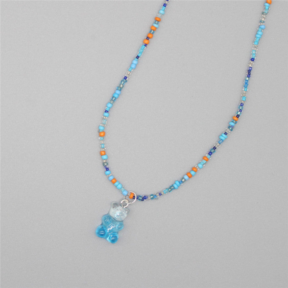 Blue-themed beaded necklace with a mix of light blue, orange, navy, and iridescent seed beads paired with a translucent blue gummy bear pendant. Playful single-strand design laid flat on a light gray background, showcasing its fresh and vibrant summer style.