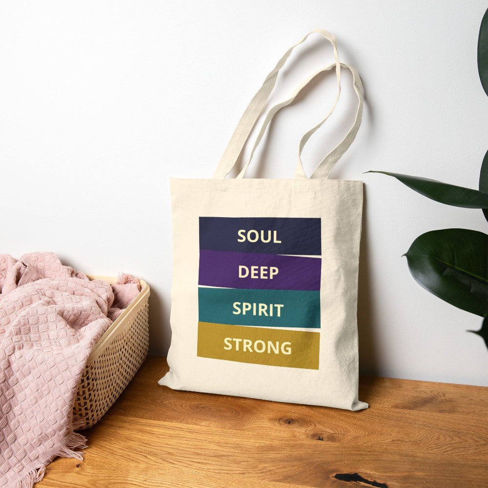 Natural canvas tote bag with "SOUL DEEP SPIRIT STRONG" motivational quote in colorful stacked blocks (navy, purple, teal, gold), styled on wooden surface with pink blanket and plant.