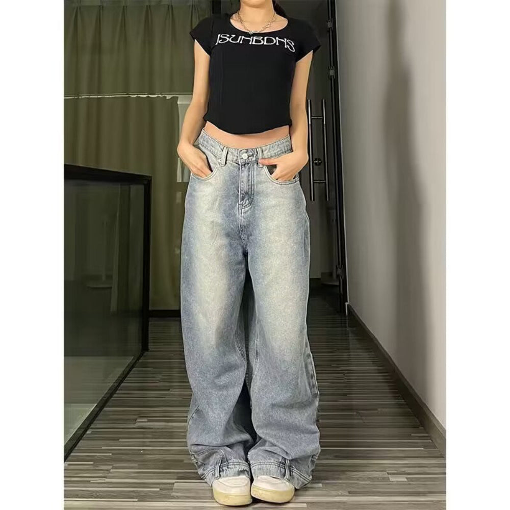 Woman modeling light-wash blue wide-leg baggy jeans with hands in front pockets, paired with a black crop top and white sneakers, standing in a hallway with wooden flooring.