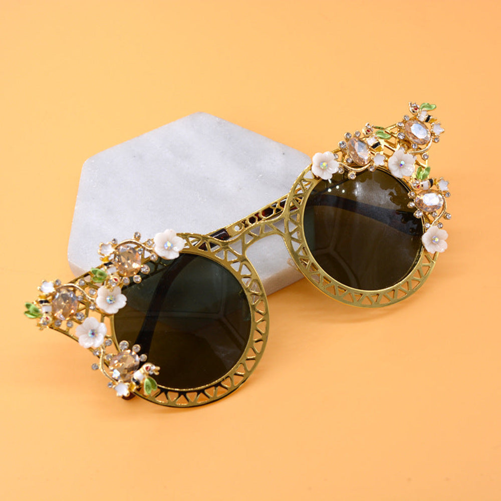 Elegant gold metal cat-eye sunglasses featuring intricate lattice frames, white floral accents, green leaves, sparkling crystals, and dark green-brown tinted lenses, displayed on marble