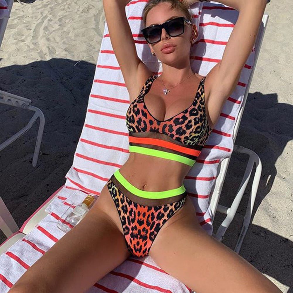 Model in leopard print neon bikini relaxing on beach lounge chair with arms raised.