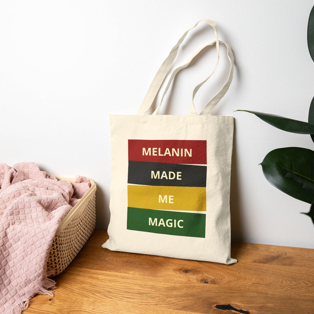 Natural canvas tote bag with "MELANIN MADE ME MAGIC" graphic on wooden table with pink throw and plant.