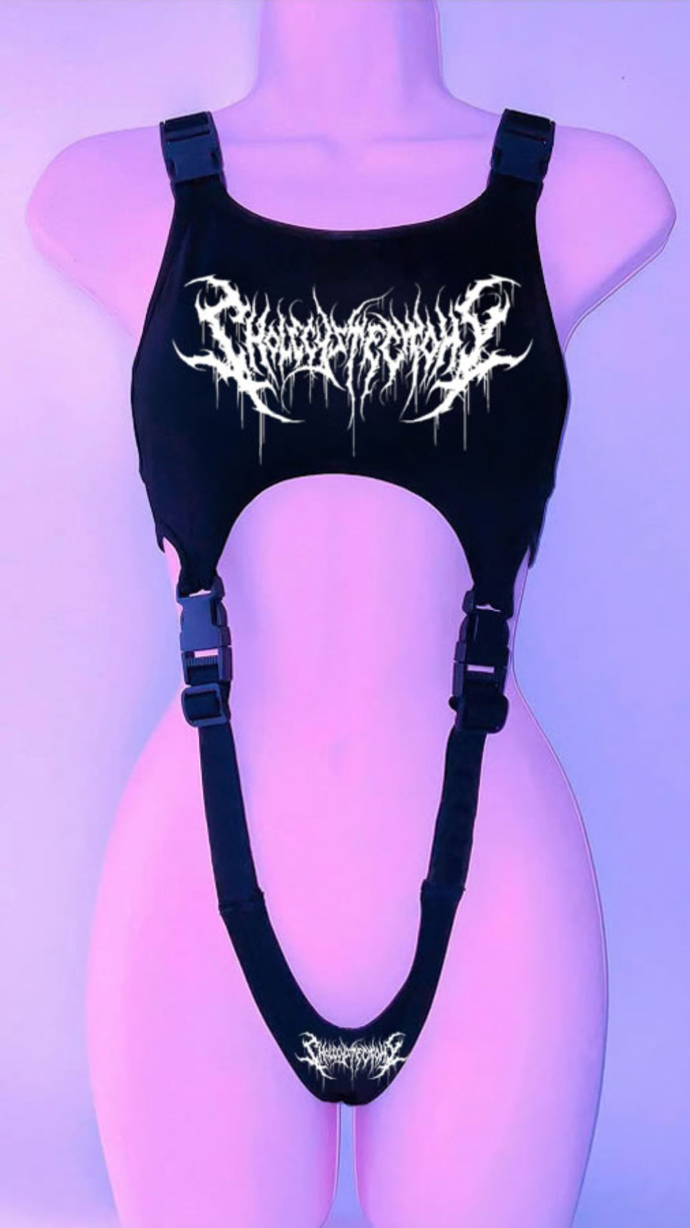Black harness-style crop top on a mannequin with adjustable buckle straps and side cut-outs. Features a bold white graphic print with skull and stylized text on the chest, plus a smaller matching graphic at the bottom, shown under purple lighting.