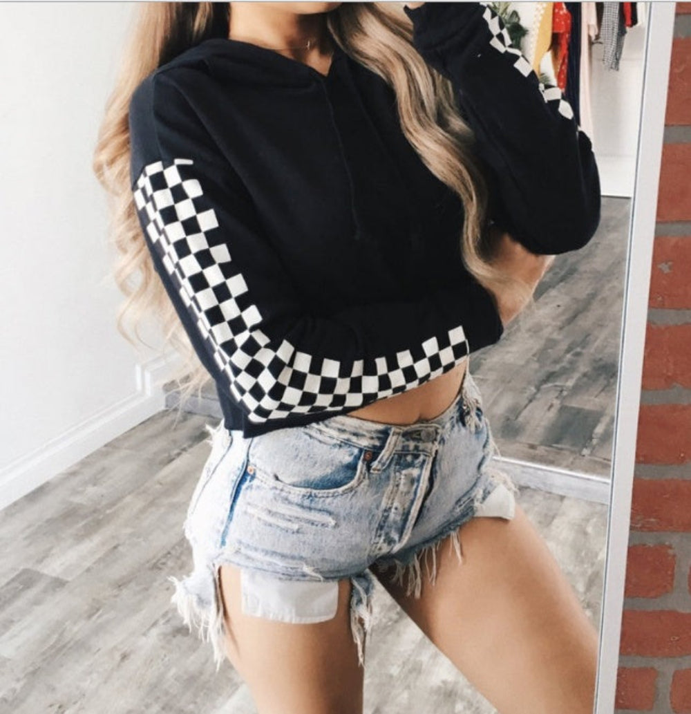 Woman with long blonde hair modeling a black cropped hoodie featuring bold black-and-white checkerboard pattern on the sleeves. Paired with light-wash distressed denim shorts with frayed hems, posing confidently in a mirror selfie.