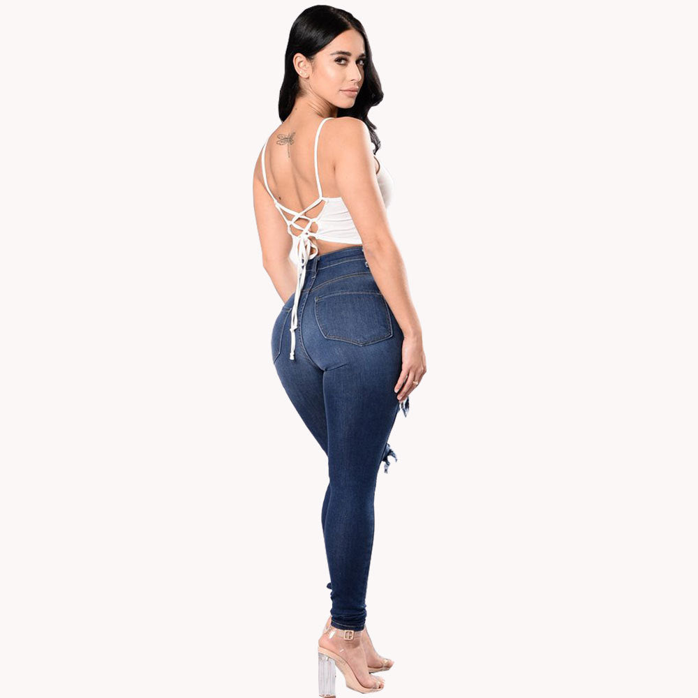 Three-quarter back view of a woman with long dark hair wearing high-waisted dark blue skinny jeans that hug her curves, featuring subtle distressing and frayed hems at the ankles. She is styled with a white strappy lace-up crop top with crisscross back ties, clear high-heeled sandals, and a small tattoo on her upper back.