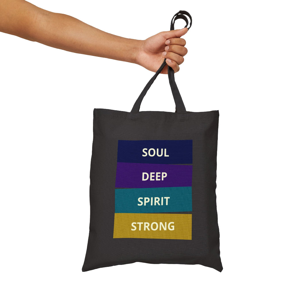 Black canvas tote bag with "SOUL DEEP SPIRIT STRONG" motivational quote in colorful stacked blocks (navy, purple, teal, gold), held by hand against white background.