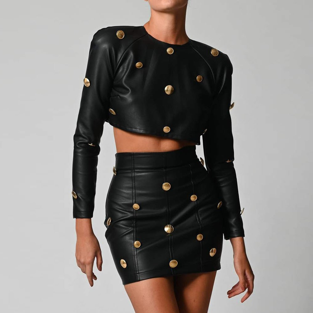 Model wearing a chic black faux leather matching outfit: cropped long-sleeve top with gold button embellishments and a fitted mini skirt with matching gold buttons. Full torso fashion pose highlighting the structured silhouette and luxurious metallic details on a clean light background.