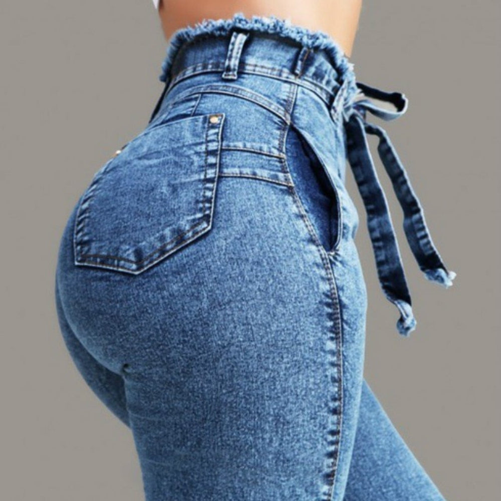 Close-up back view of woman wearing blue high-waisted skinny jeans with self-tie waist detail, paperbag waist, and frayed raw edges. Studio fashion shot highlighting the trendy design for Boutique Jolie.