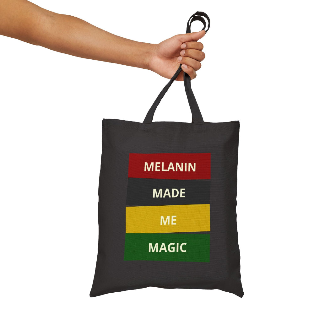 Hand holding black canvas tote bag with "MELANIN MADE ME MAGIC" graphic in red, black, yellow, and green blocks.