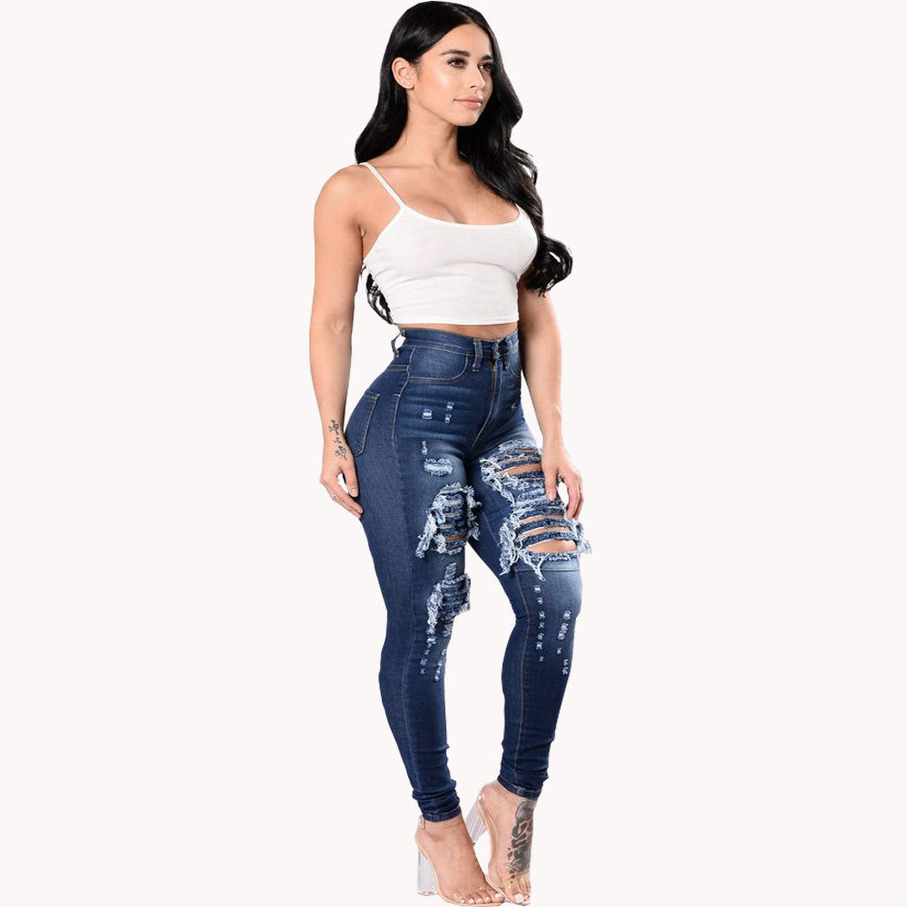Front view of a woman with long dark wavy hair wearing high-waisted dark blue skinny jeans with heavy distressing, large ripped holes, and frayed details on the thighs and knees. Styled with a white spaghetti strap crop top and clear high-heeled sandals, highlighting tattoos on her arm and lower leg.