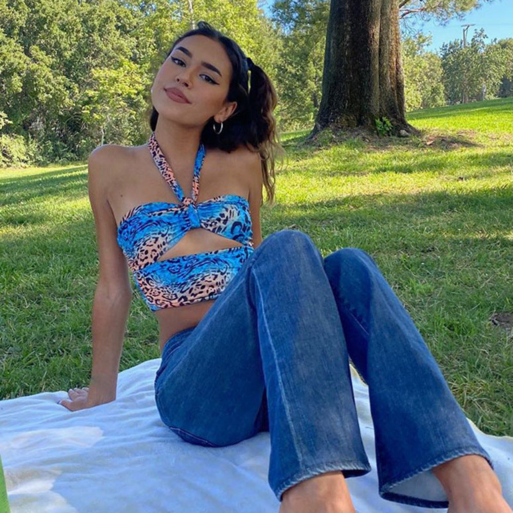 Woman wearing a blue leopard print halter crop top with twist-front detail and cut-out design, paired with wide-leg blue denim jeans. Photographed outdoors sitting on a white blanket in a sunny park setting with green grass and trees.