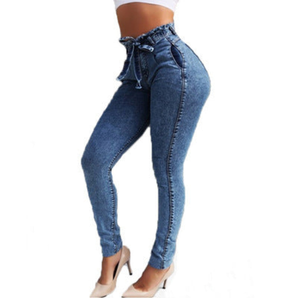 Woman modeling light blue high-waisted skinny jeans with self-tie waist detail and paperbag waist, paired with a white crop top and nude high heels. Side view studio fashion shot for Boutique Jolie.