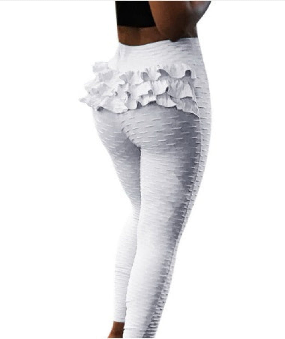 Model wearing white stretch high-waisted leggings featuring a textured crinkle fabric and eye-catching tiered ruffle layers around the hips and waist. Rear side view highlighting the flattering fit and decorative ruffle design on a plain white background.