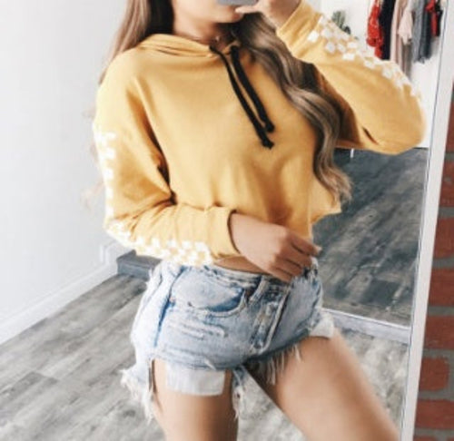 Woman with long wavy hair modeling a mustard yellow cropped hoodie with white checkered pattern on the sleeves and black drawstrings. Paired with light-wash distressed denim shorts, posing in a mirror selfie.