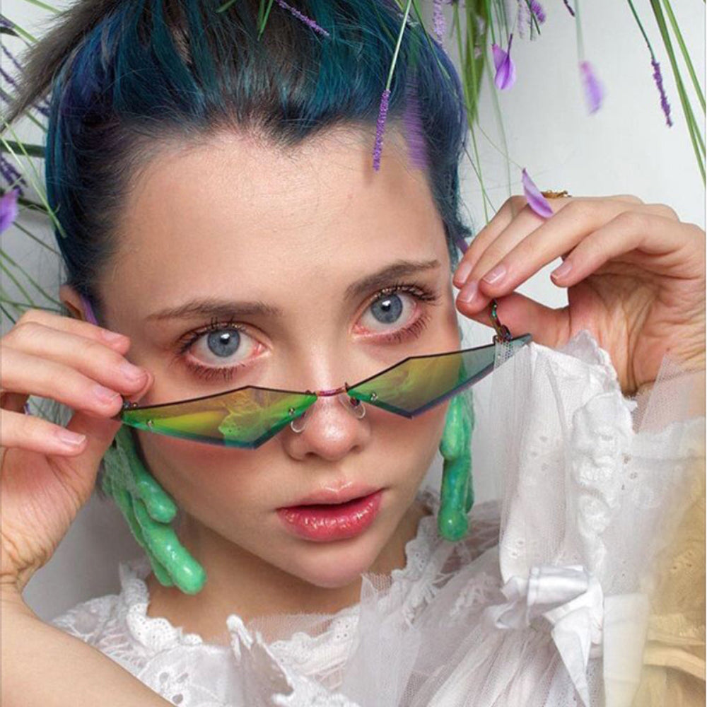 Woman with teal hair wearing rimless triangle sunglasses with green-gold lenses.