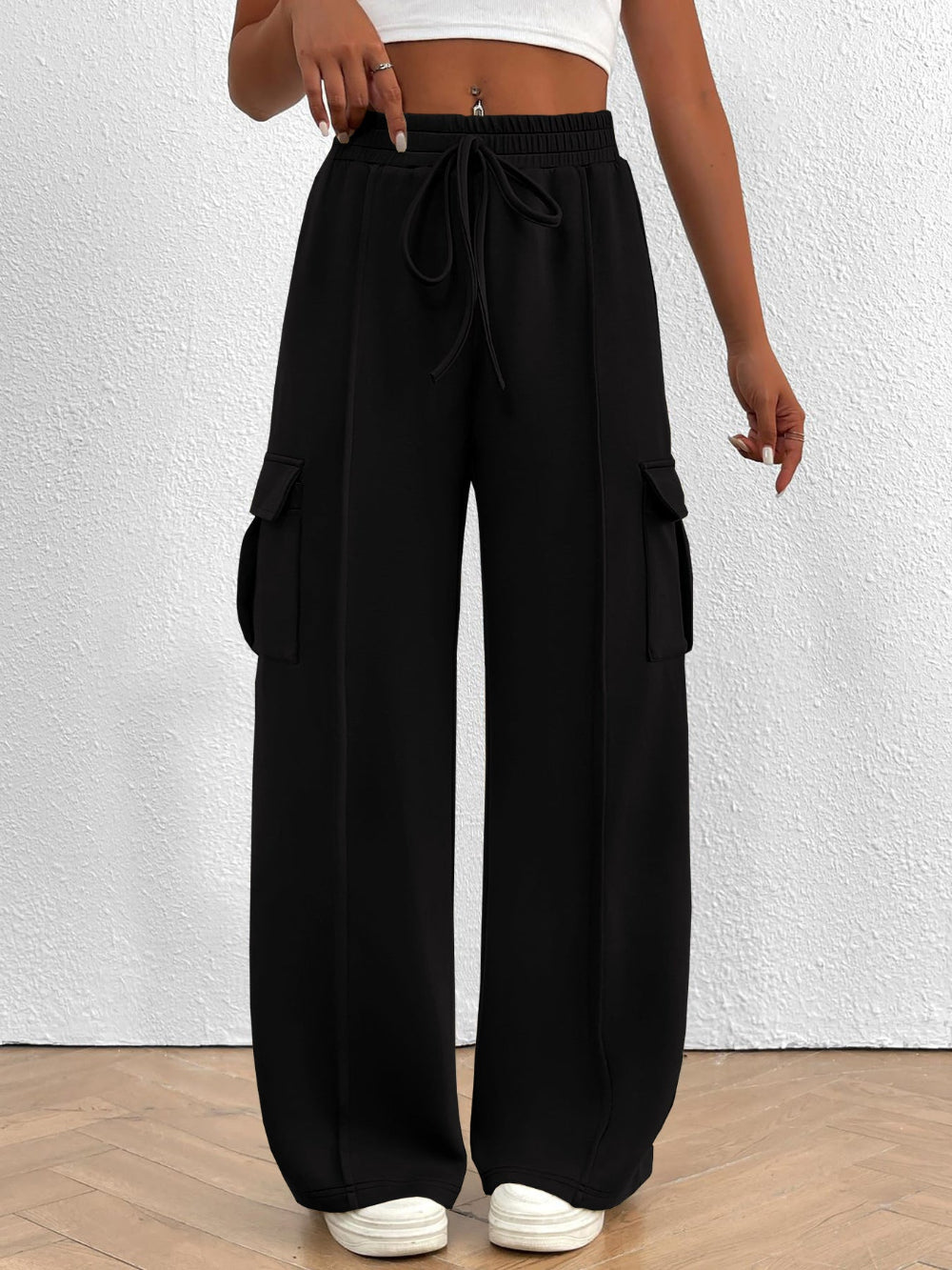 Woman wearing black wide leg cargo pants with drawstring waist and large side pockets paired with a white top and white sneakers against a textured white wall background