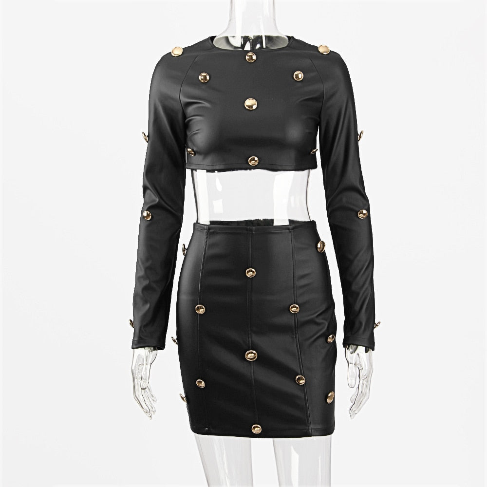 Black faux leather two-piece outfit on a mannequin: cropped long-sleeve top and fitted mini skirt, both featuring prominent gold button embellishments in a military-inspired style. Full product view highlighting the structured silhouette and metallic details on a plain white background.