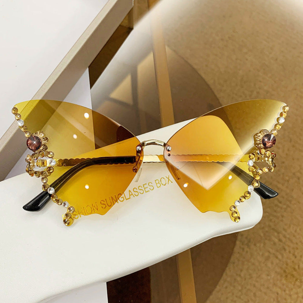 Rimless butterfly sunglasses with yellow-amber lenses and gold crystal wing details, displayed on a white glasses case.