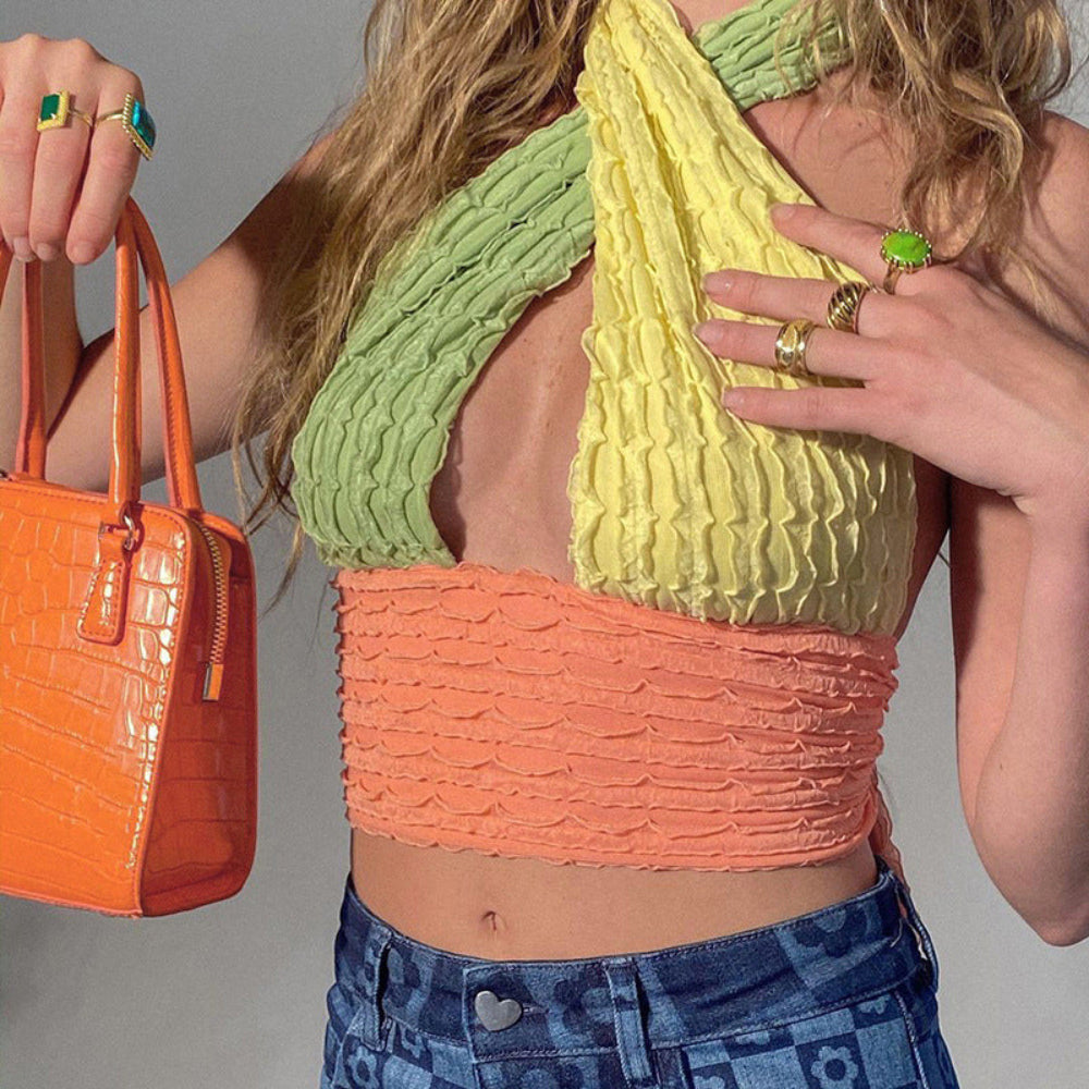 Woman with long wavy blonde hair modeling a vibrant crinkled halter crop top in lime green, yellow, and coral pink with textured pleated fabric and scalloped edges. The backless design features a dramatic twisted halter neck. Styled with blue patterned jeans and holding a bright orange crocodile-embossed handbag, accessorized with bold colorful rings.