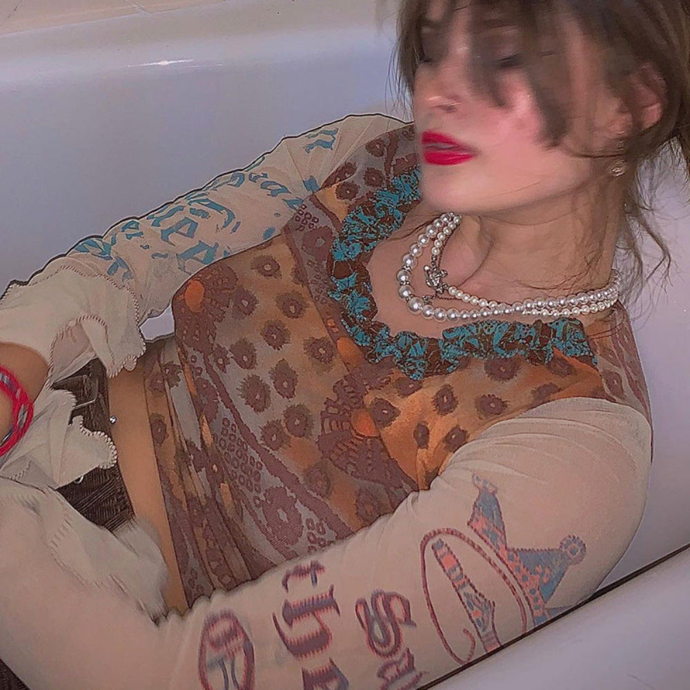 Woman with brown hair and bold red lipstick reclining in a white bathtub, wearing a sheer long-sleeve mesh top with intricate brown, orange, and turquoise paisley-style patterns and graphic prints. The top features distressed frayed sleeve details, layered with multiple pearl necklaces and a visible belly button piercing.