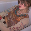 Woman with brown hair and bold red lipstick reclining in a white bathtub, wearing a sheer long-sleeve mesh top with intricate brown, orange, and turquoise paisley-style patterns and graphic prints. The top features distressed frayed sleeve details, layered with multiple pearl necklaces and a visible belly button piercing.