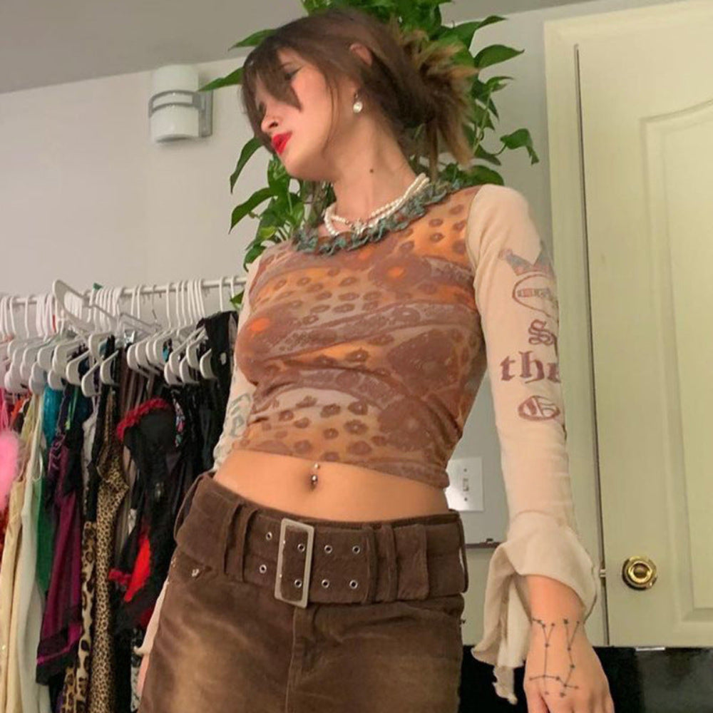 Woman with brown hair in a high ponytail wearing a sheer long-sleeve mesh crop top featuring intricate brown, orange, and turquoise paisley patterns. Styled with a wide brown belt over brown pants, layered pearl necklace, and visible belly button piercing, posing indoors near a clothing rack.