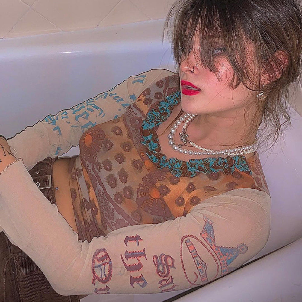 Woman with brown hair and red lipstick reclining in a white bathtub, wearing a sheer long-sleeve mesh crop top with intricate brown, orange, and turquoise paisley patterns plus graphic prints on the sleeves. Styled with multiple layered pearl necklaces and a brown belt, creating a stylish bohemian look.