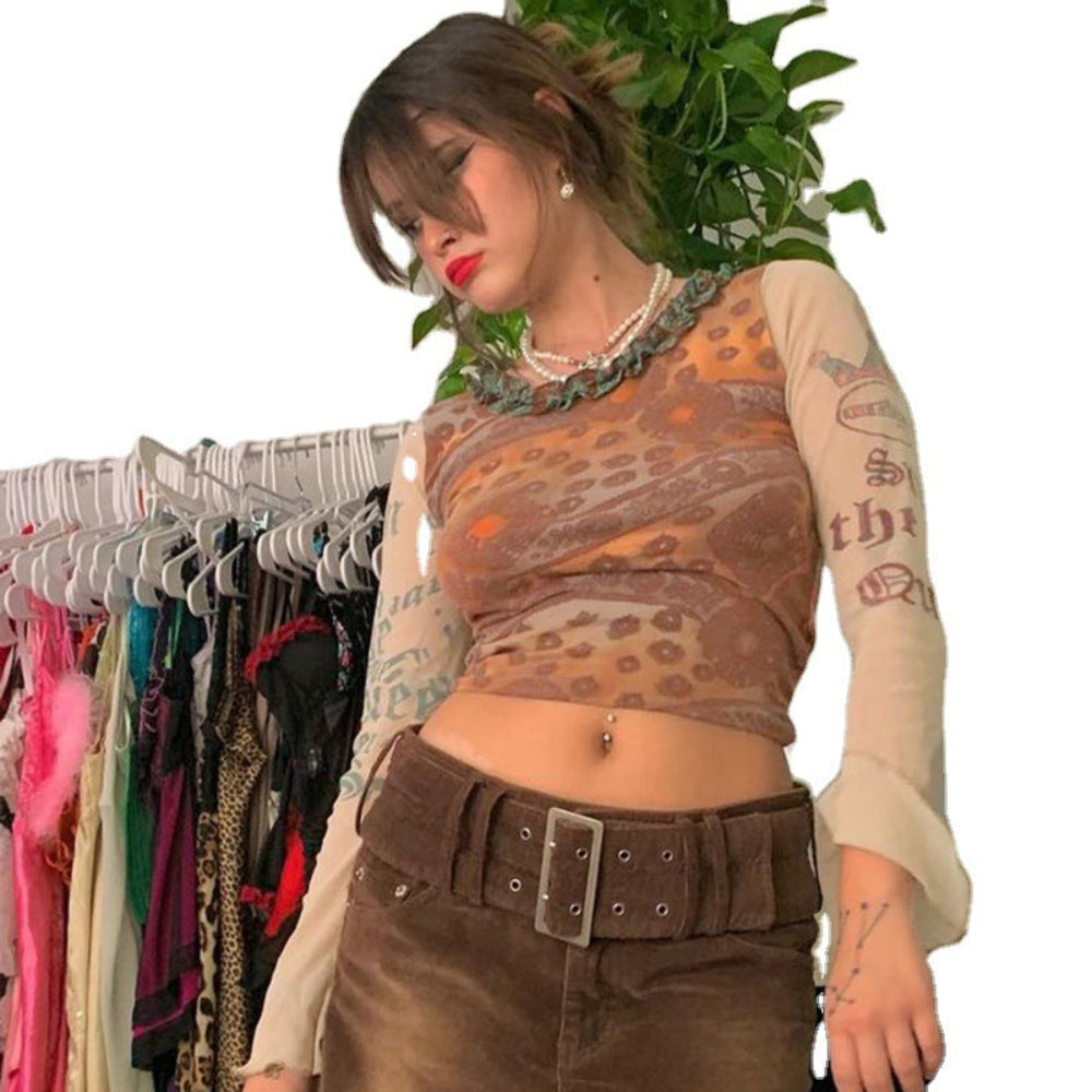 Woman with brown hair and bangs wearing a sheer long-sleeve mesh crop top with intricate brown, orange, and turquoise paisley patterns over cream sleeves featuring graphic prints. Styled with a wide brown belt with large silver buckle over brown pants, layered pearl necklace, and visible belly button piercing, posing indoors near a clothing rack.