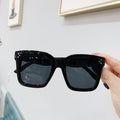 Black oversized square sunglasses with dark lenses and crystal accents at the temples, held in hand.