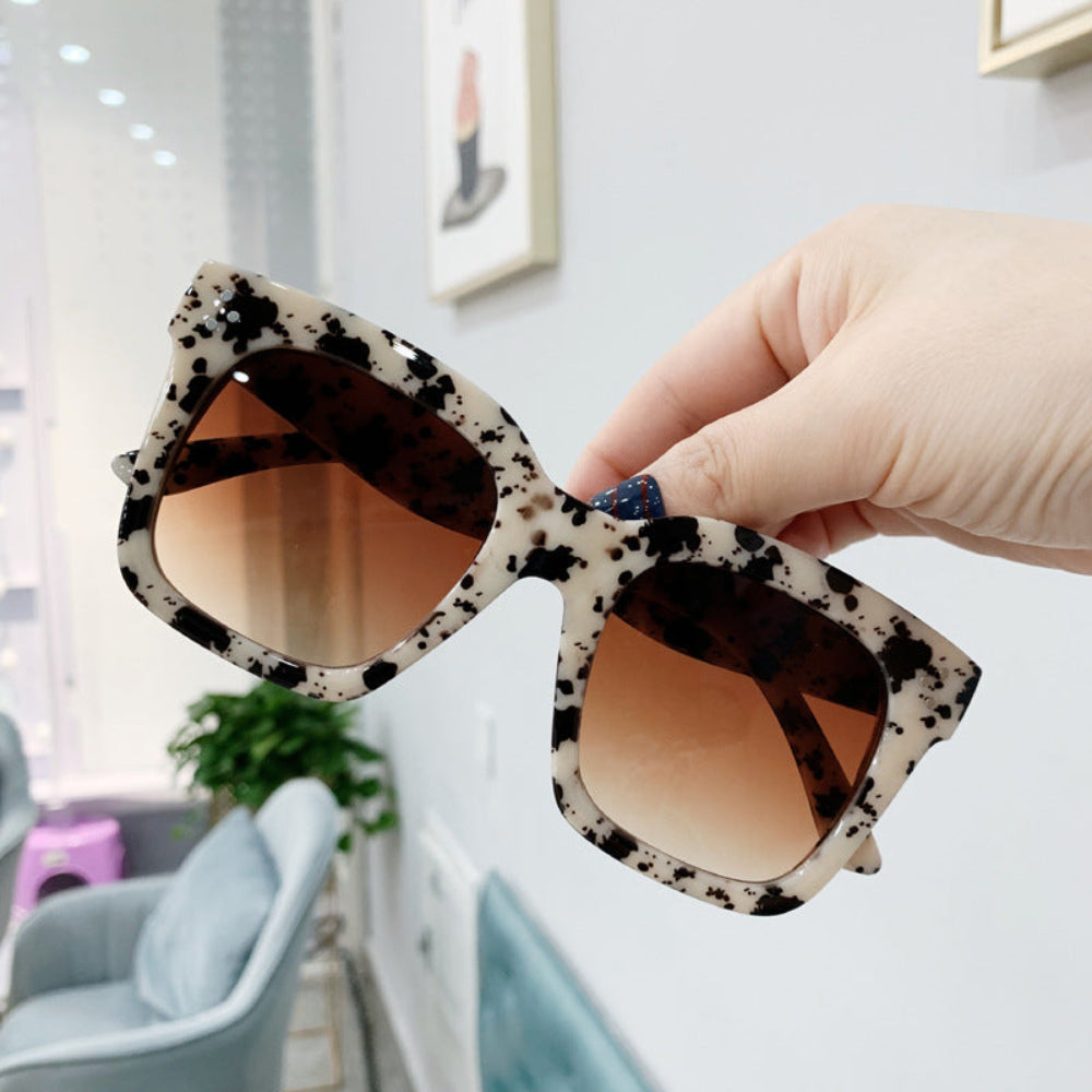 Cream and black splatter oversized square sunglasses with brown gradient lenses, held in hand.