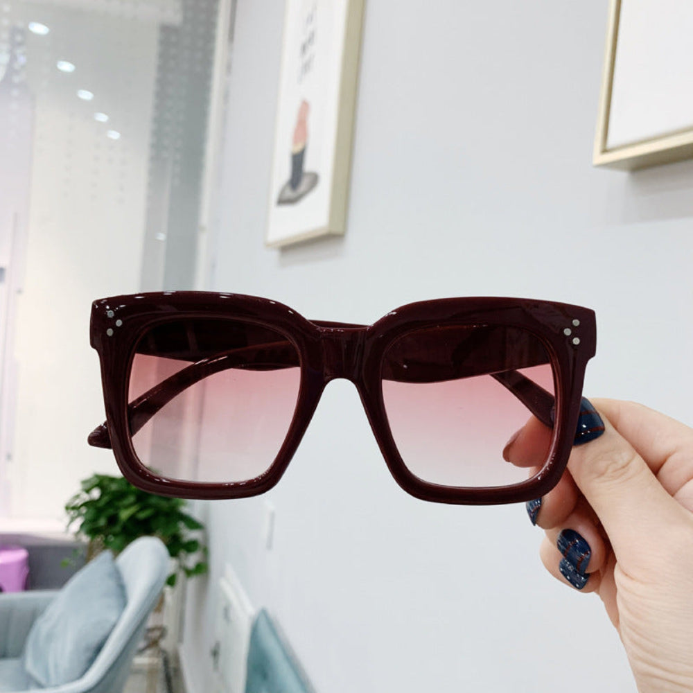 Burgundy oversized square sunglasses with pink gradient lenses and crystal temple accents, held in hand.