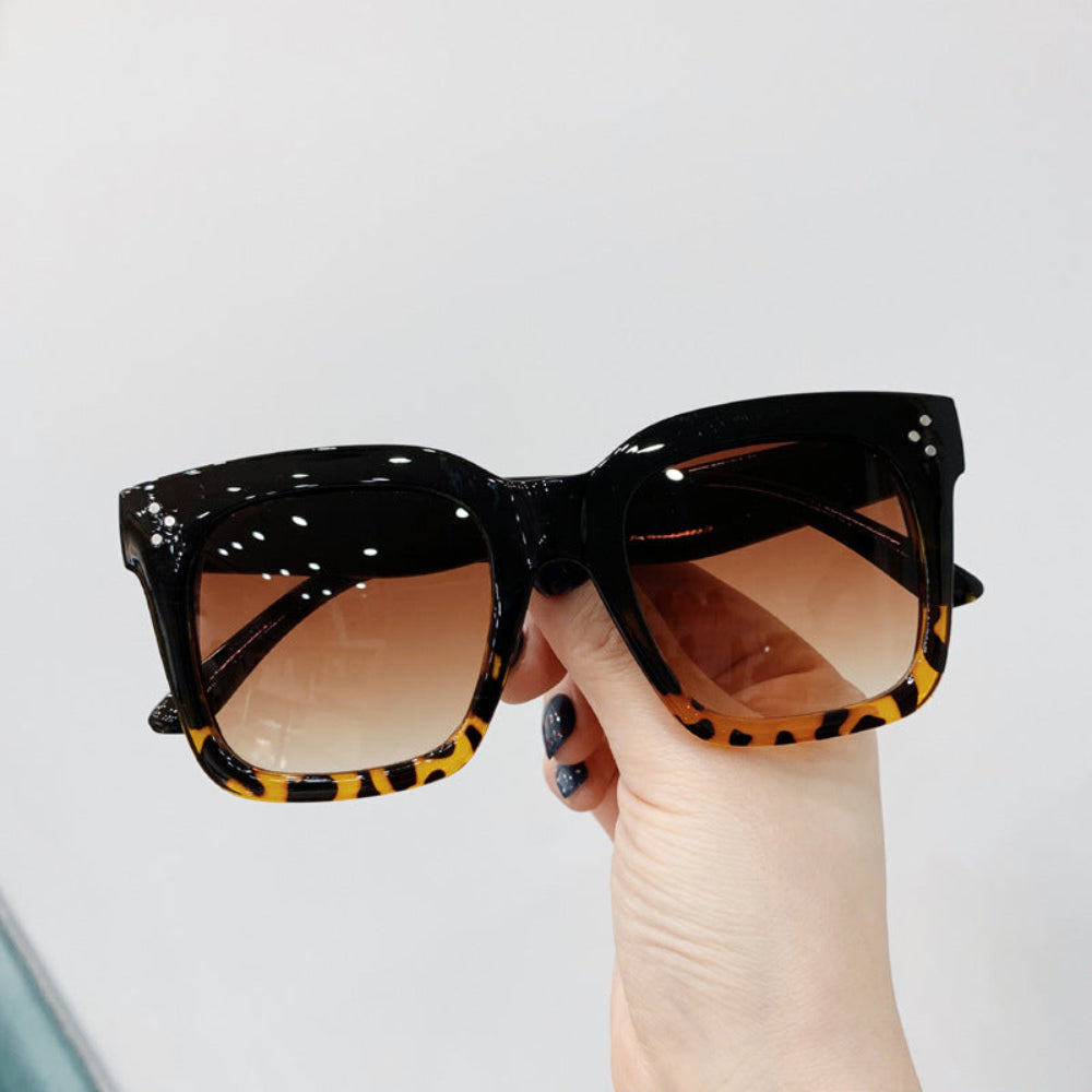 Black oversized square sunglasses with leopard-print lower rims and brown gradient lenses, held in hand.