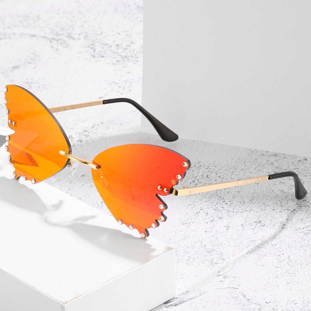 Rimless butterfly sunglasses with orange lenses, rhinestone edges, and gold temples, styled on a white marble surface.