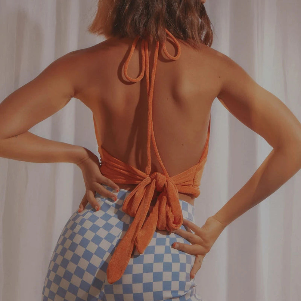 Orange velvet halter crop top with open back, long tie closures, and ruched detailing, shown from the back on a model wearing checkered pants.