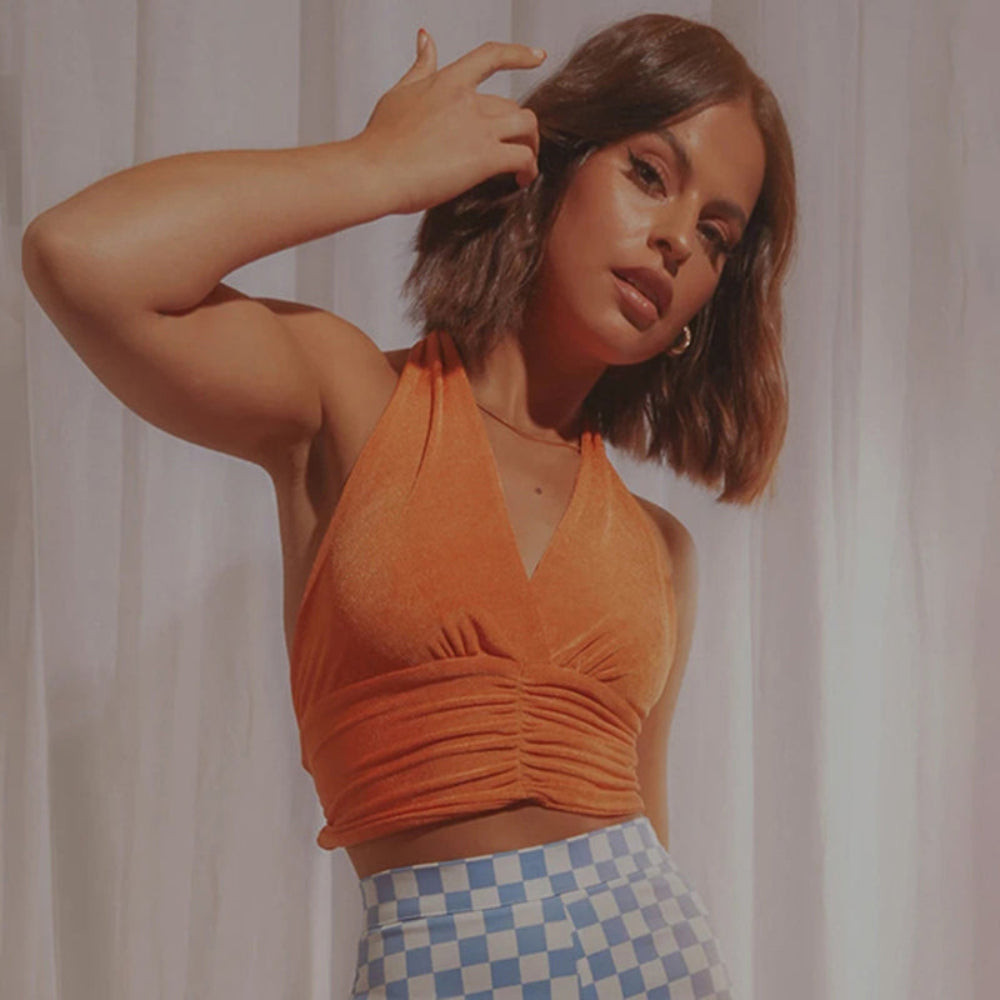 Orange velvet halter crop top with deep V-neck and ruched waist detail, worn by a model with checkered pants.