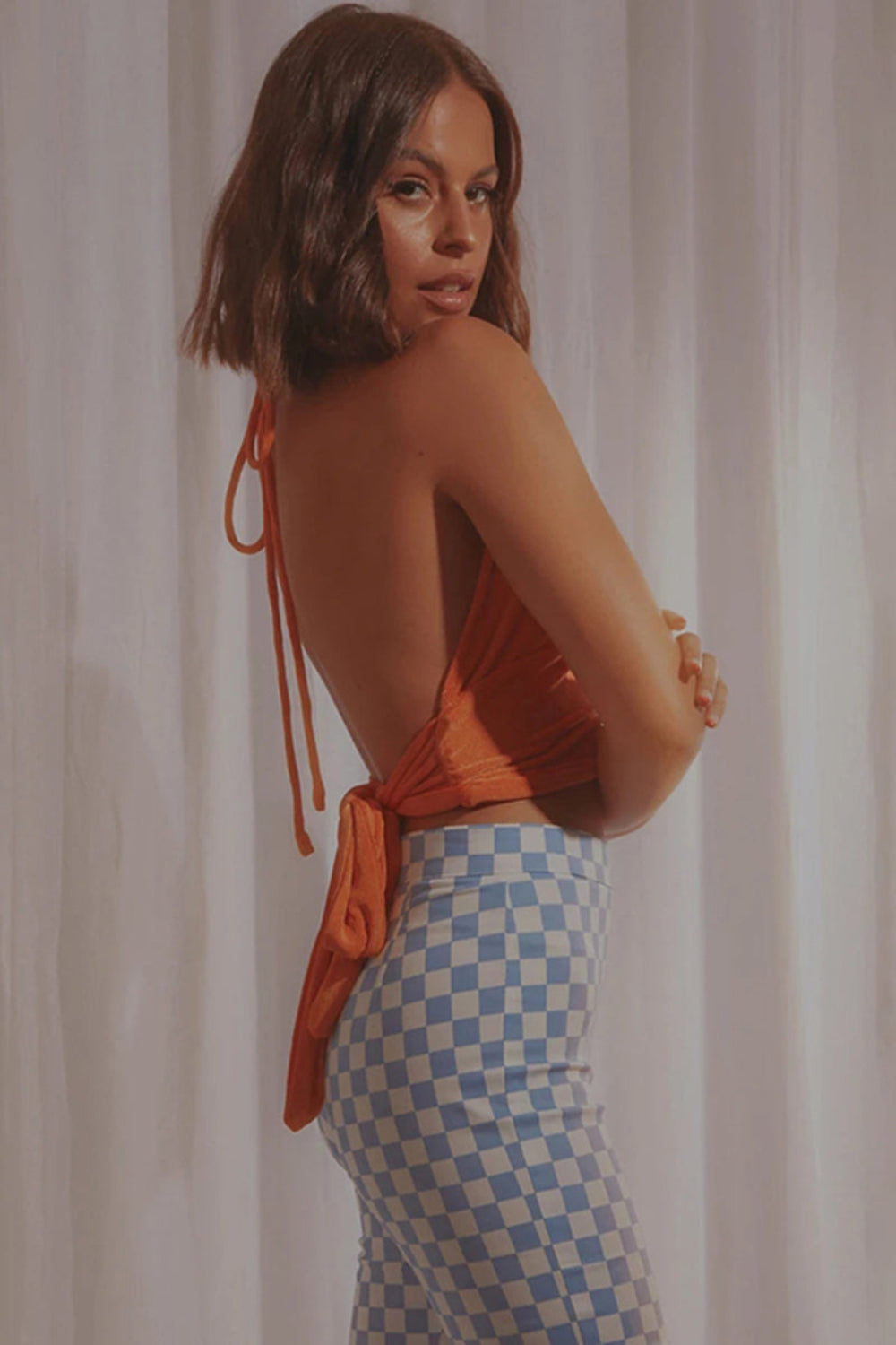 Orange velvet halter crop top with tie-back closures and ruched detailing, shown from the back on a model wearing checkered pants.