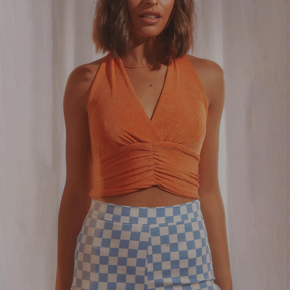 Orange velvet halter crop top with deep V-neck and ruched waist detail, paired with blue and white checkered pants, worn by a model.