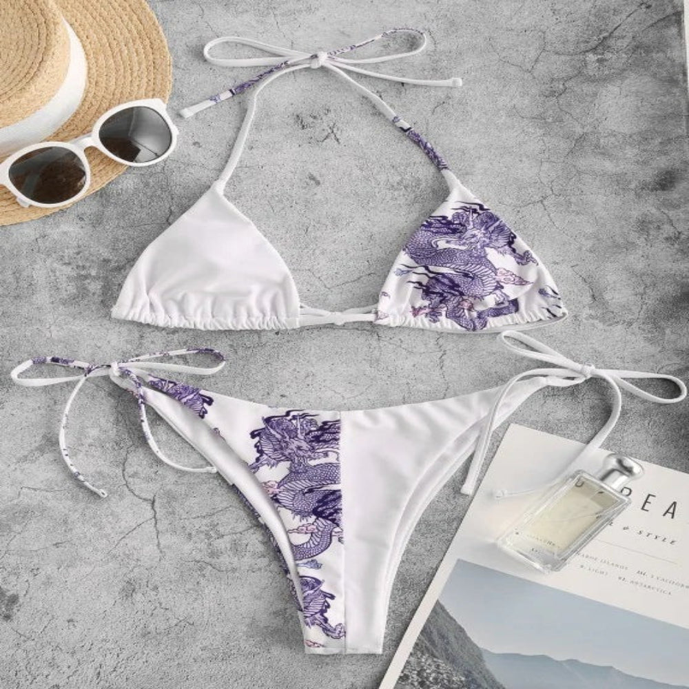 White bikini with purple floral pattern on a gray surface with sunglasses and a hat.