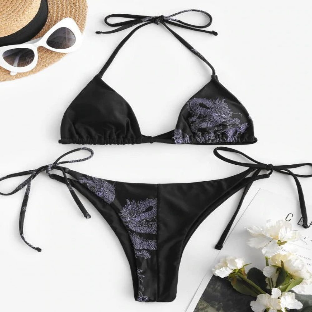 Black bikini set with floral patterns on a white background, accompanied by sunglasses and flowers.