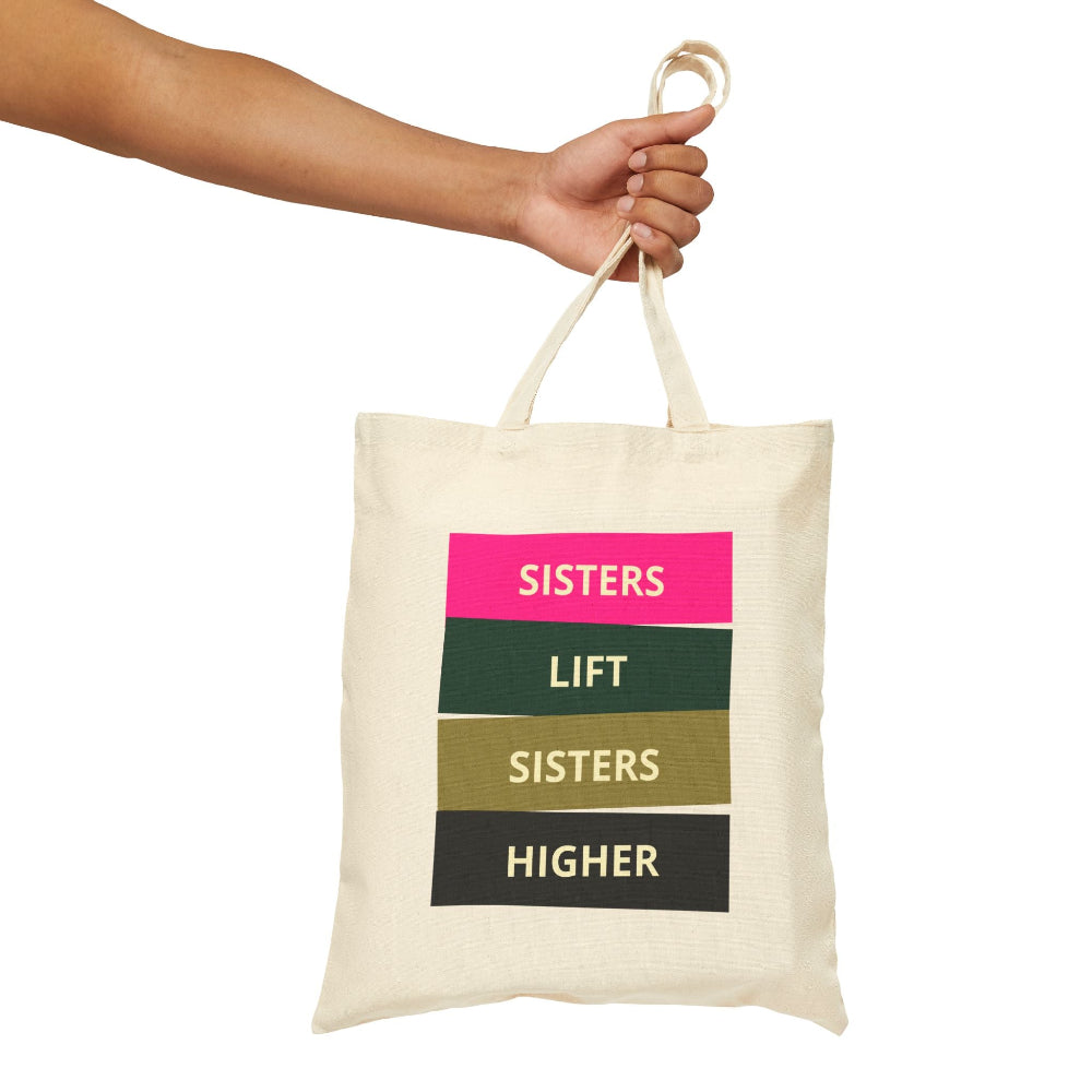 Hand holding natural canvas tote bag with "SISTERS LIFT SISTERS HIGHER" colorful block text.