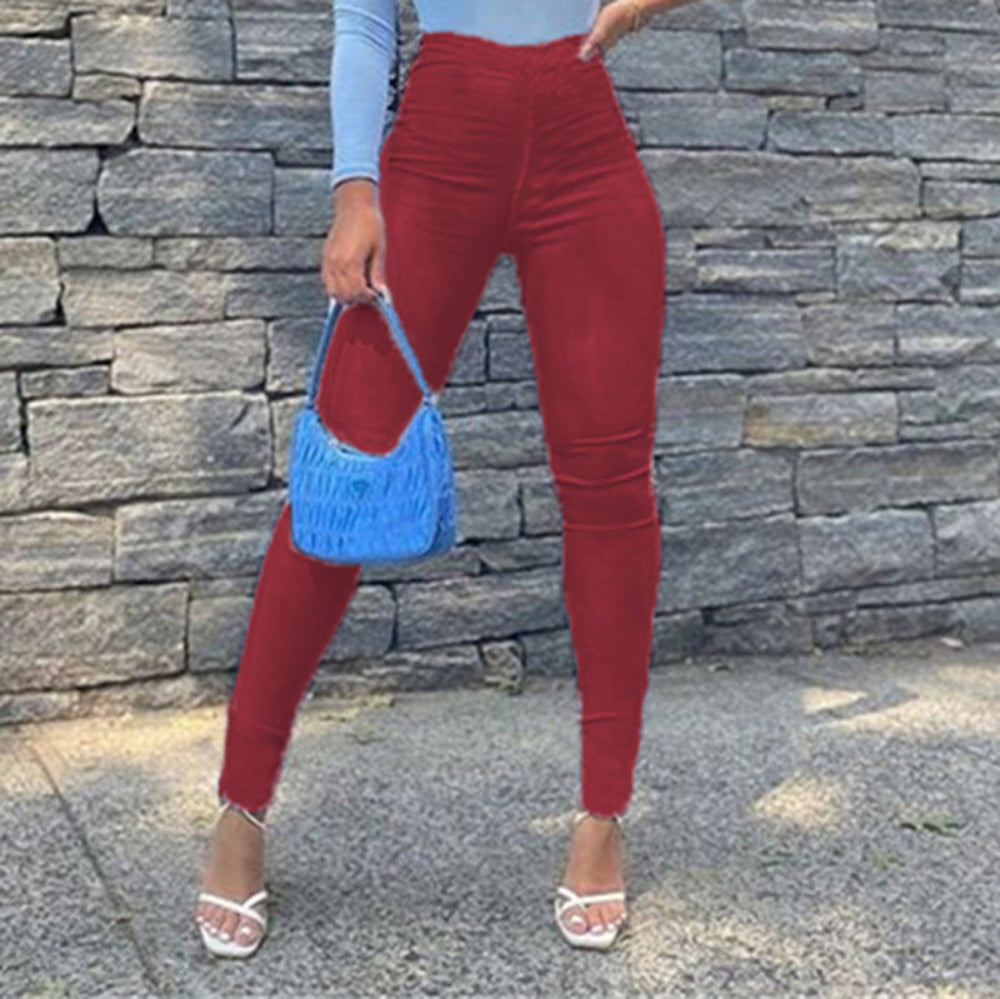 Woman wearing high-waisted red skinny pants and a light blue long-sleeve top, holding a bright blue woven handbag and posing in white strappy heels against a stone wall.